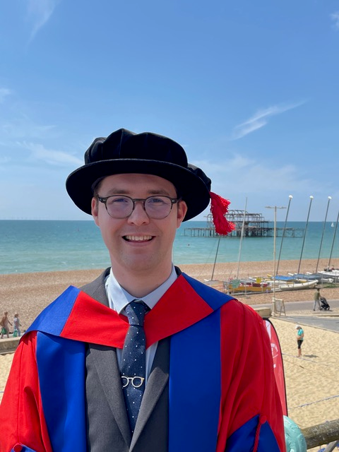 Dr. Tim Galsworthy, shown in academic regalia on a beach with blue ocean in the background.